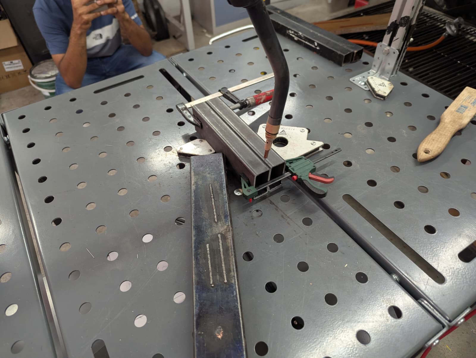 The MIG torch held at a constant standoff above a clamped steel box section, positioned on the seam before the arc is armed.