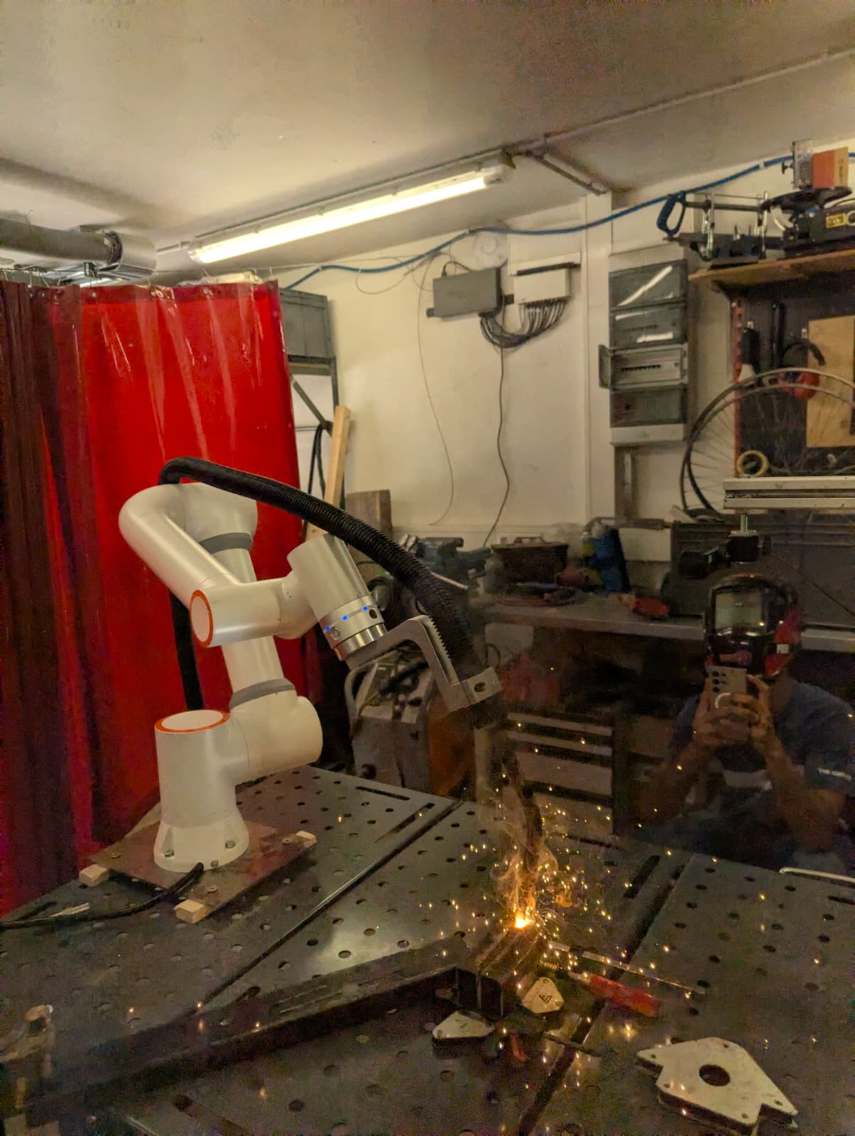 Omnicron's arm mounted to a welding table, torch down on a clamped joint with the arc struck and sparks flying, screen curtain behind.