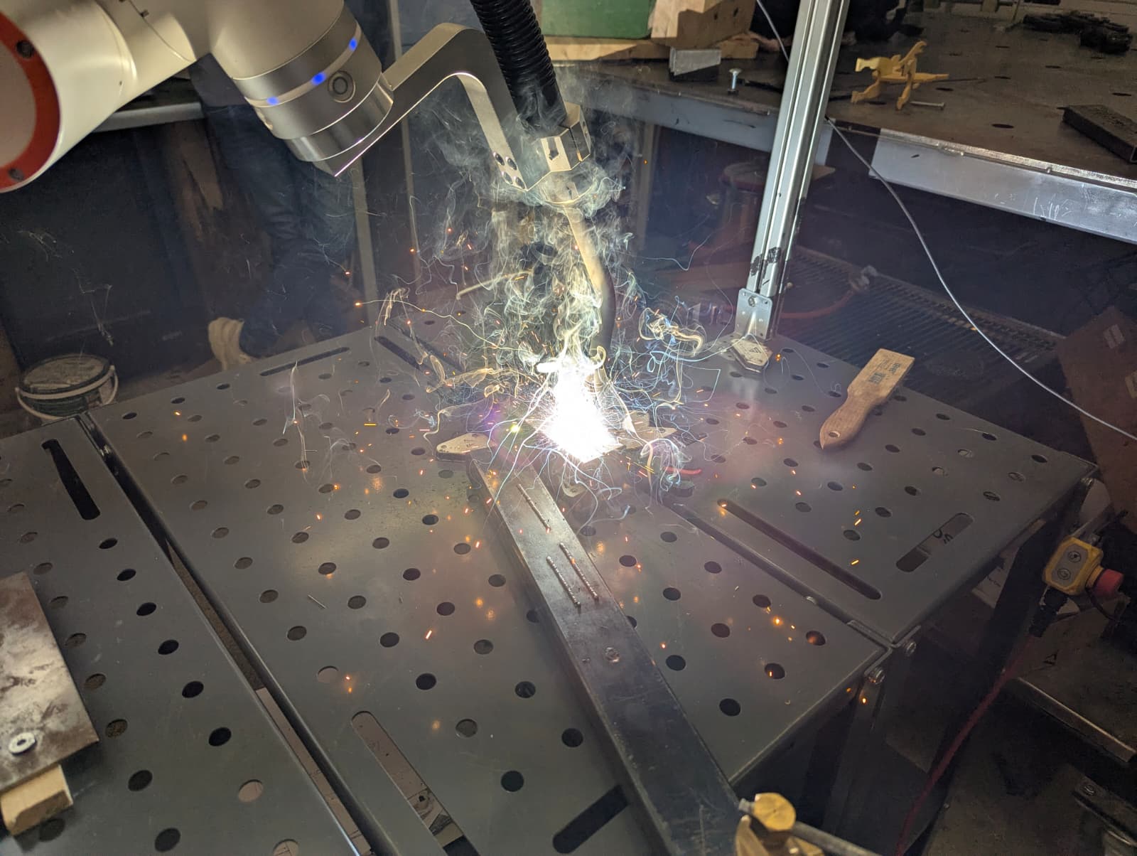 Close view through the welding screen of the torch mid-pass, arc bright at the joint with spatter arcing off the table.